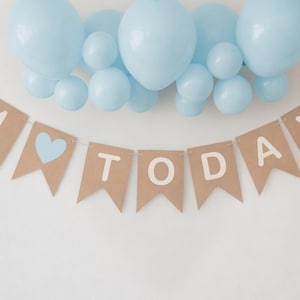 Op de afbeelding: Een feestelijke banner met de woorden "1 TODAY" in witte letters, een blauw hart en een cluster van lichtblauwe ballonnen. De banner is gemaakt van bruin papier en is perfect voor een eerste verjaardag.