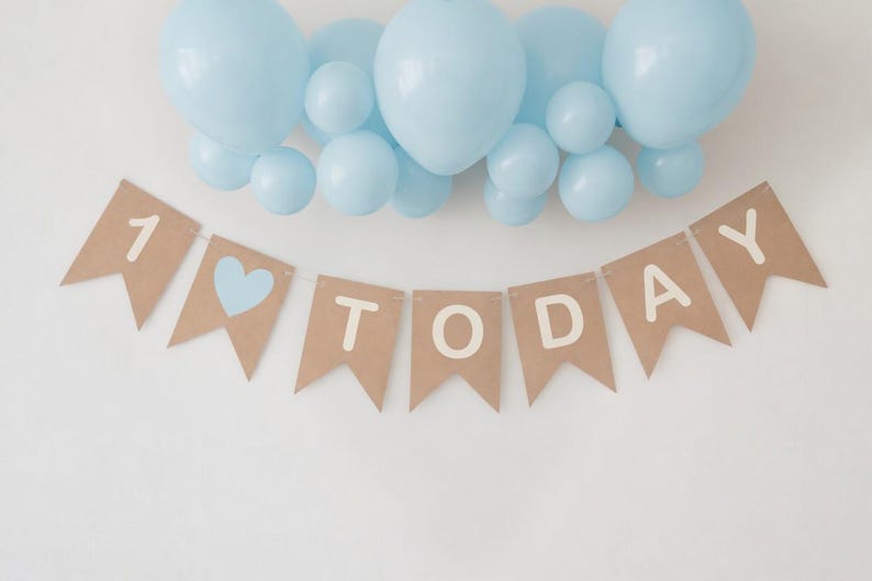 Op de afbeelding: Een feestelijke banner met de woorden "1 TODAY" in witte letters, een blauw hart en een cluster van lichtblauwe ballonnen. De banner is gemaakt van bruin papier en is perfect voor een eerste verjaardag.