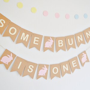May include: A festive banner with the words "SOME BUNNY IS ONE" in white letters on brown pennants. Pink bunny silhouettes and pastel polka dots add to the celebratory theme. Perfect for a first birthday party.