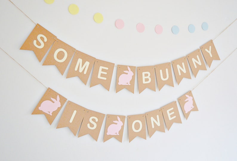May include: A festive banner with the words "SOME BUNNY IS ONE" in white letters on brown pennants. Pink bunny silhouettes and pastel polka dots add to the celebratory theme. Perfect for a first birthday party.