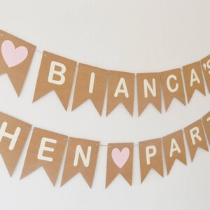 May include: A brown paper banner with white letters spelling "Bianca's Hen Party" with pink hearts between the words.