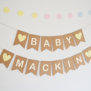 Puede incluir: Una pancarta de papel marr&oacute;n con el texto "BABY MACKIN" en letras blancas y corazones amarillos. La pancarta est&aacute; atada con un cordel y cuelga de una pared blanca.
