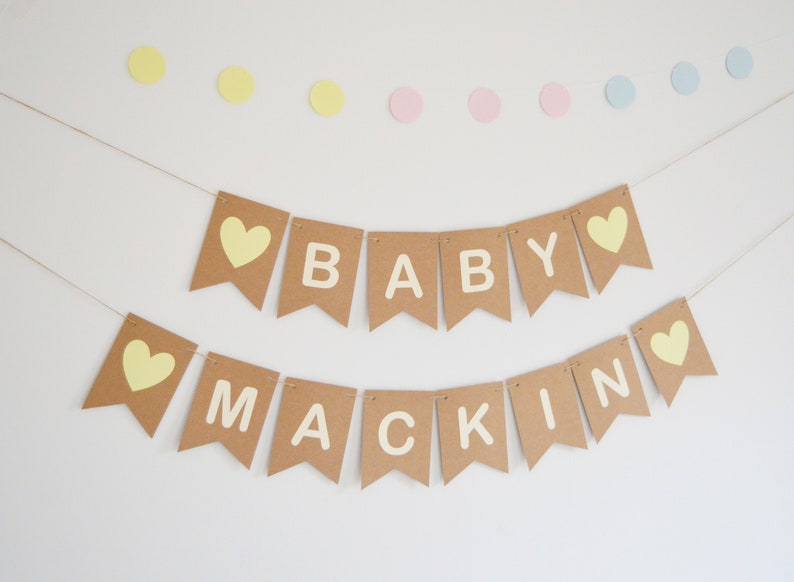 Puede incluir: Una pancarta de papel marr&oacute;n con el texto "BABY MACKIN" en letras blancas y corazones amarillos. La pancarta est&aacute; atada con un cordel y cuelga de una pared blanca.
