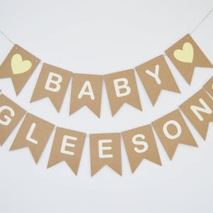 Puede incluir: Una pancarta de papel marr&oacute;n con las palabras "Baby Gleeson" en letras blancas y corazones amarillos. La pancarta est&aacute; enhebrada con un cordel.