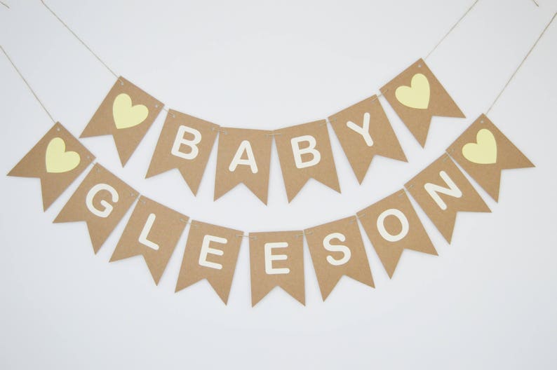 Puede incluir: Una pancarta de papel marr&oacute;n con las palabras "Baby Gleeson" en letras blancas y corazones amarillos. La pancarta est&aacute; enhebrada con un cordel.
