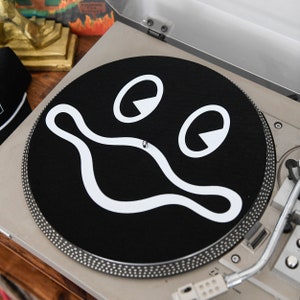 Puede incluir: Una alfombrilla de tocadiscos negra con un diseño de cara sonriente blanca. La cara tiene dos ojos grandes y una boca ancha y curvada.