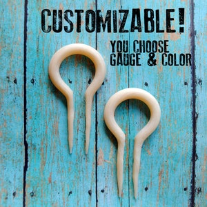 May include: Two white bone ear gauges, shaped like a teardrop with a loop at the top. The text "CUSTOMIZABLE! YOU CHOOSE GAUGE & COLOR" is above the gauges.