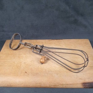 Op de afbeelding: Een vintage metalen garde met een houten handvat. De garde heeft een lang, gebogen handvat en meerdere draadlussen om eieren te kloppen.