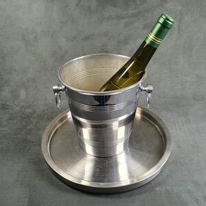 May include: A silver ice bucket with a bottle of white wine inside. The bucket has a handle on each side and sits on a silver tray.