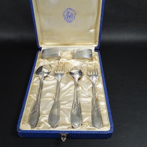May include: A blue velvet box with a satin lining contains a set of silver cutlery. The cutlery includes four pieces: two forks, a spoon, and a knife. The box is closed and the cutlery is visible through the clear plastic lid.
