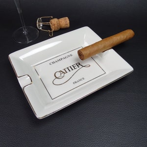 Könnte beinhalten: Weiße Aschenbecher aus Keramik mit goldener Einfassung und dem Aufdruck "Champagne Cartier France". Ein brauner Zigarre liegt auf dem Aschenbecher.