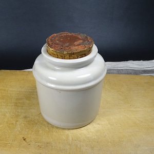 Puede incluir: Tarro de cerámica blanco con tapa de corcho. El tarro tiene un cuerpo redondeado y un labio ligeramente acampanado. La tapa de corcho es marrón y tiene una textura ligeramente rugosa.