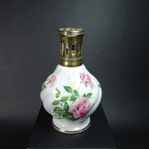 May include: A white ceramic oil lamp with a gold-coloured metal top and base. The lamp is decorated with pink roses and green leaves. The lamp has a bulbous shape and sits on a small, round base.