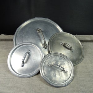 Könnte beinhalten: Vier silberne Aluminium-Topfdeckel in verschiedenen Größen mit Griffen. Die Deckel sind übereinander auf einer hellgrauen Stoffoberfläche gestapelt.