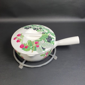 May include: White ceramic saucepan with a lid, featuring a colorful botanical design of raspberries and blackberries. The saucepan has a long, white handle and sits on a silver-coloured metal trivet. The lid has a white handle for easy removal.