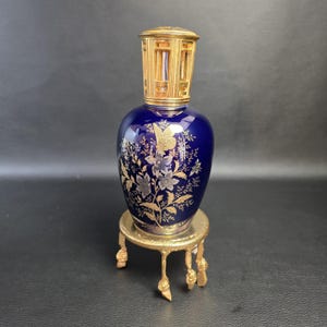 May include: A deep blue glass fragrance lamp featuring a gold-toned metal crown top and a gold-toned metal stand with four legs. The body of the lamp is decorated with a gold and silver floral pattern that includes a butterfly and various blossoms.