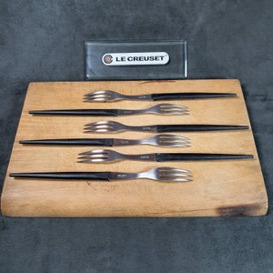 Può includere: Sei forchette Le Creuset con manici neri sono disposte su un tagliere di legno. Le forchette hanno un design elegante e moderno con rebbi in acciaio inossidabile. Sullo sfondo è presente una custodia rettangolare trasparente con il logo Le Creuset.