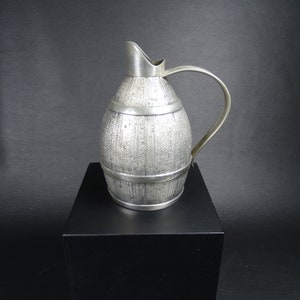 May include: A silver pitcher shaped like a barrel with a textured surface. The pitcher is sitting on a black pedestal.