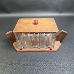 Puede incluir: Un recipiente vintage de madera y vidrio con tapa. El recipiente tiene forma rectangular con base y tapa de madera. La tapa presenta un pequeño pomo redondo rojo. La parte de vidrio tiene un diseño texturizado.