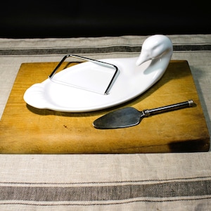 Puede incluir: Tabla de queso de cerámica blanca con forma de ganso con un cortador de queso de alambre y un cuchillo de queso plateado sobre una tabla de cortar de madera.