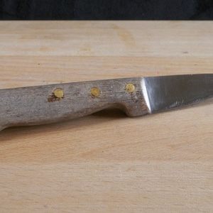 Peut inclure: Un petit couteau avec un manche en bois et une lame en métal. Le manche est orné de rivets en laiton. Le couteau est posé sur une surface en bois.