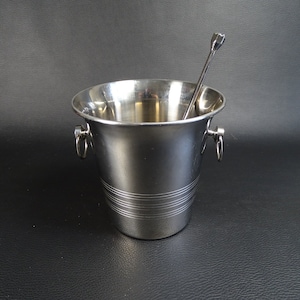 Puede incluir: Un cubo de hielo de plata con una pinza de hielo de plata. El cubo tiene un fondo redondeado y un borde alrededor de la parte superior. La pinza tiene un mango con un bucle en el extremo.