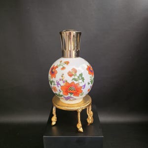 Op de afbeelding: Een bolvormige witte keramische geurlamp met een kleurrijk bloemenpatroon met oranje, rode, paarse en groene details. De lamp is voorzien van een zilverkleurige geventileerde dop en staat op een goudkleurige metalen standaard met drie poten.