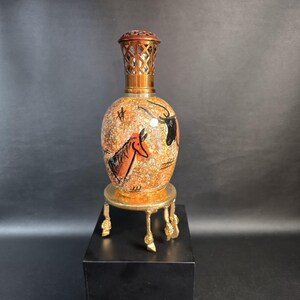 May include: A ceramic fragrance lamp with a speckled orange and yellow finish, featuring black hand-painted animal figures. The lamp has a metallic bronze-colored lattice top and rests on a gold-toned metal stand with four decorative legs.