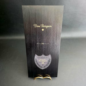 Könnte beinhalten: Eine schwarze Dom Pérignon Champagnerbox mit goldfarbenen Buchstaben und einem goldenen Ständer. Die Box hat eine strukturierte Oberfläche und zeigt das Dom Pérignon-Logo sowie die Wörter "P2" und "2000".