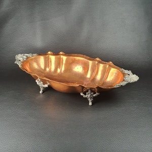 May include: An oval copper serving bowl with three decorative feet. The bowl has a shiny, polished finish and is accented with silver-toned details.