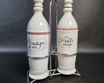 May include: Two white ceramic cruets with matching stoppers held in a chrome wire caddy. The left bottle features black script text reading Vinaigre de vin, and the right bottle features black script text reading Huile vierge, both with thin orange bands.
