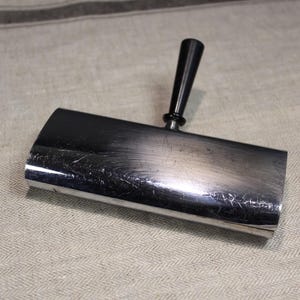Peut inclure: Un cendrier en métal argenté et noir avec une poignée noire. Le cendrier est rectangulaire et a une surface lisse.