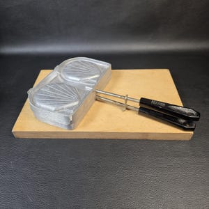 Könnte beinhalten: Ein silberfarbenes Waffeleisen aus Metall mit zwei muschelförmigen Formen. Das Gerät verfügt über zwei lange Metallgriffe mit schwarzen Kunststoffgriffen, die jeweils mit dem Text "ALFA" und "MADE IN ITALY" versehen sind. Es liegt auf einem Holzbrett.