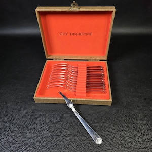 Pode incluir: Um conjunto de garfos para marisco em metal prateado numa caixa retangular bege com forro interior laranja brilhante. A marca GUY DEGRENNE está impressa a preto na tampa interior. Um garfo está fora da caixa e apresenta um motivo de concha.