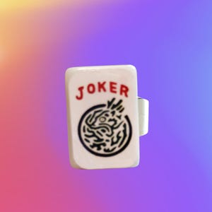 May include: A white rectangular ring featuring the word JOKER printed in red letters above a black circular graphic design. The ring has a smooth, glossy finish and a simple band structure visible on the side, presenting a clear and distinct visual.