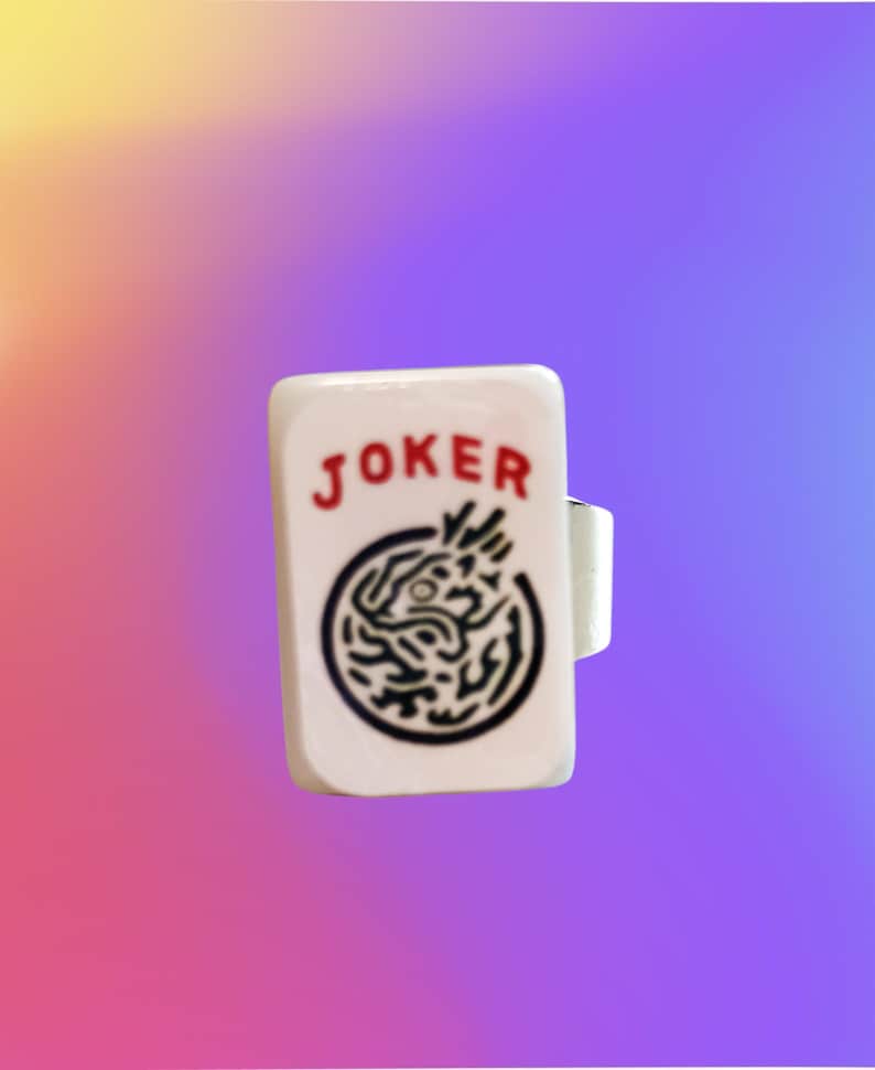 May include: A white rectangular ring featuring the word JOKER printed in red letters above a black circular graphic design. The ring has a smooth, glossy finish and a simple band structure visible on the side, presenting a clear and distinct visual.