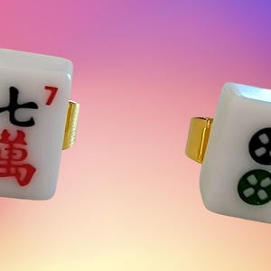 May include: Two rings featuring white rectangular mahjong tiles as the centerpiece. One tile displays the black Chinese character for seven and a red number seven, while the other shows two circular symbols in black and green with a red number two.