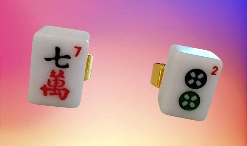 May include: Two rings featuring white rectangular mahjong tiles as the centerpiece. One tile displays the black Chinese character for seven and a red number seven, while the other shows two circular symbols in black and green with a red number two.