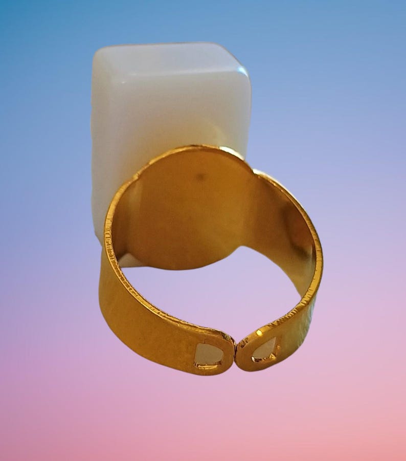 May include: A gold-toned adjustable ring featuring a smooth, rectangular, opaque white stone setting. The metal band has a polished finish and an open design at the back, allowing for size adjustments to fit various finger widths comfortably.