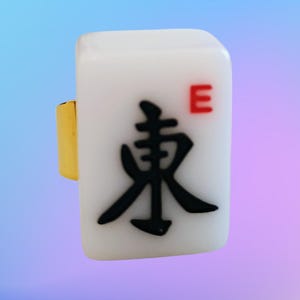 May include: A ring featuring a rectangular white mahjong tile with a gold-colored metal band. The tile displays a large black Chinese character for East and a small red letter E in the upper right corner, set against a smooth, glossy surface.