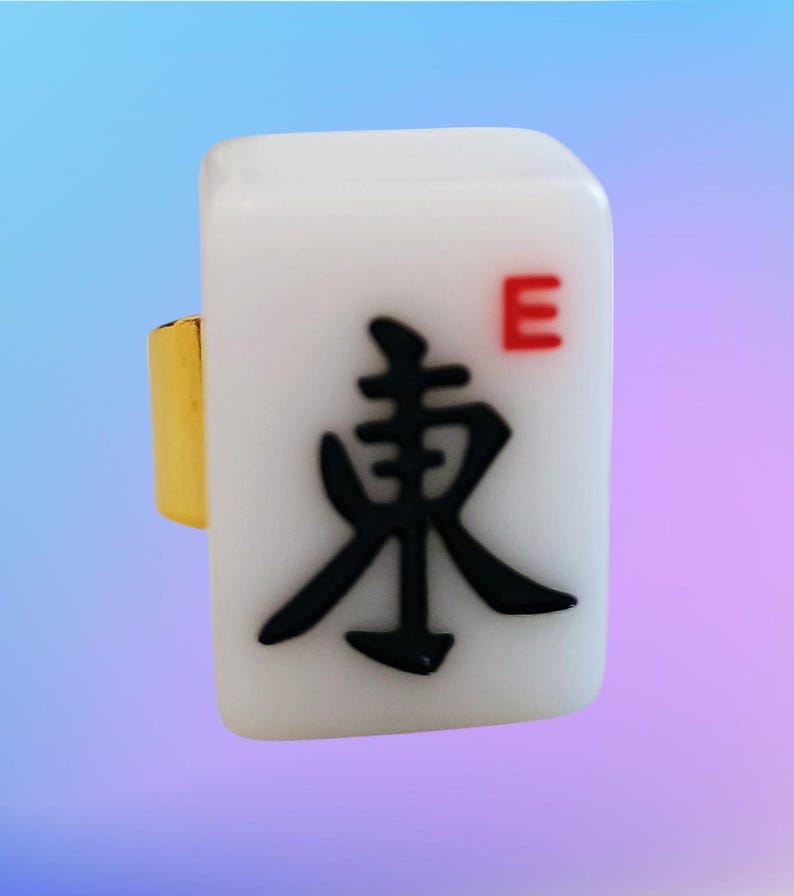 May include: A ring featuring a rectangular white mahjong tile with a gold-colored metal band. The tile displays a large black Chinese character for East and a small red letter E in the upper right corner, set against a smooth, glossy surface.