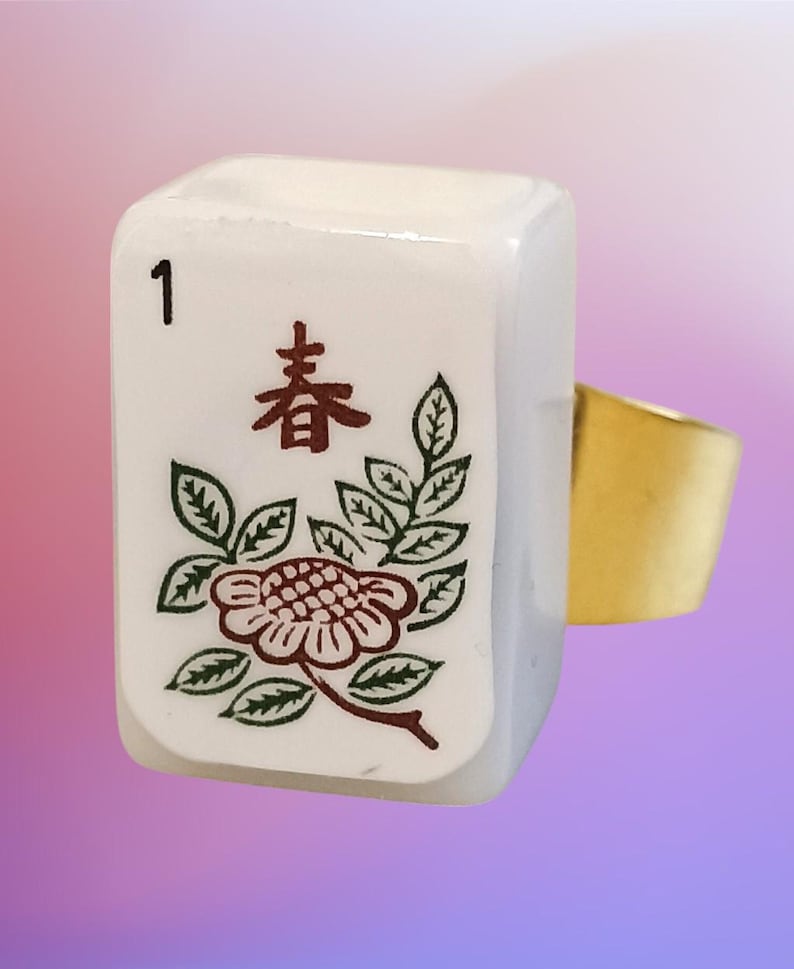 May include: A ring featuring a rectangular white mahjong tile as the centerpiece. The tile displays the number 1 in black, a brown Chinese character, and a green and brown floral illustration. The ring band is made of a smooth, polished gold-tone metal.
