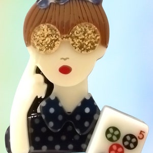 May include: A whimsical brooch featuring a stylized woman with brown hair, wearing gold glitter sunglasses and a navy blue polka dot dress. She holds a Mahjong tile with the number 5 and flower symbols. The brooch is made of glossy acrylic.