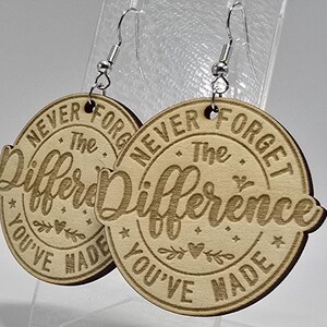 May include: A pair of wooden earrings with a circular design. The earrings feature the engraved text "NEVER FORGET The Difference YOU'VE MADE" in a decorative font. The earrings are light brown and have silver-colored hooks.