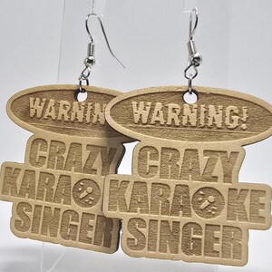 Puede incluir: Dos pendientes de madera con ganchos plateados. Cada pendiente tiene una parte superior ovalada con la palabra "WARNING" y una parte inferior rectangular con "CRAZY KARAOKE SINGER" en letras negritas. Un icono de micrófono está en el centro del rectángulo inferior.