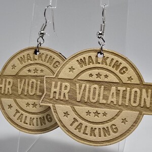 May include: A pair of wooden earrings with a stamped design. Each earring is round with the words "WALKING," "HR VIOLATION," and "TALKING" engraved. The earrings have silver-colored fish hook ear wires.