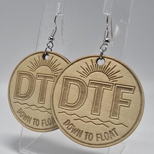 May include: Two wooden disc earrings with silver-coloured hooks. Each earring has the letters "DTF" above the words "Down to Float" and a sun design. The earrings are circular in shape and made of light-coloured wood.