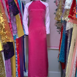 May include: A pink long-sleeved dress with white sleeves and a mandarin collar. The dress is on a mannequin in front of a rack of colourful clothing.