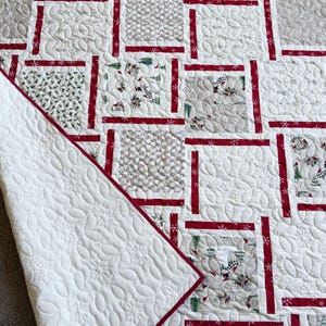May include: A white and red Christmas quilt with a patchwork design. The quilt features squares with various patterns, including snowflakes and holiday-themed prints. The quilt's border is red, and the backing is white with a snowflake pattern.