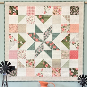 May include: A quilt with a starburst pattern in shades of green, pink, and white. The quilt is made of various floral fabrics and is hanging on a wall. The quilt is surrounded by a light green border.